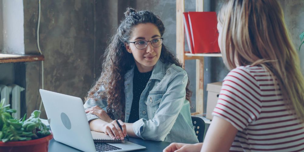 Dos mujeres conversando en una mesa con un computador portátil y un cuaderno.