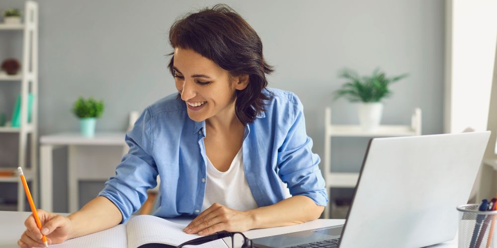 Mujer tomando apuntes en un cuaderno junto a un computador portátil.
