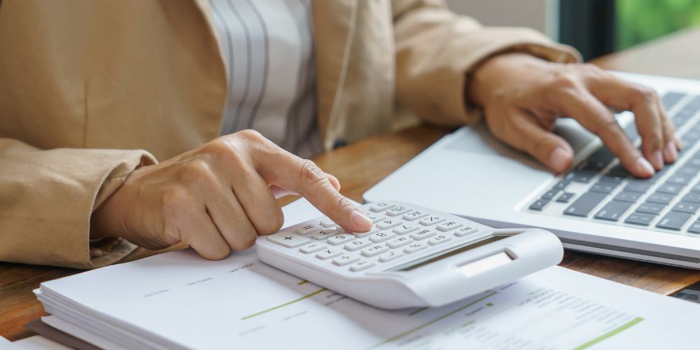Persona utilizando una calculadora junto a documentos y un computador portátil.