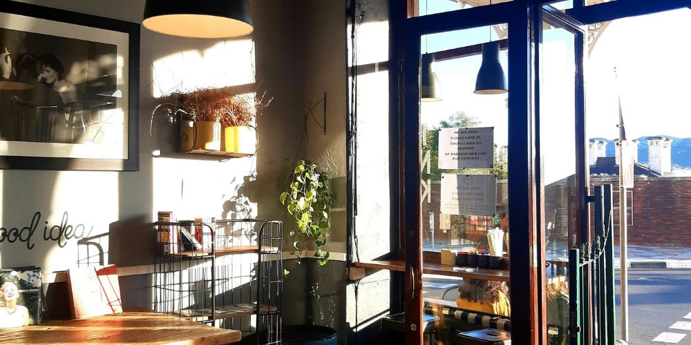 Interior de una cafetería con mesas, plantas y entrada acristalada