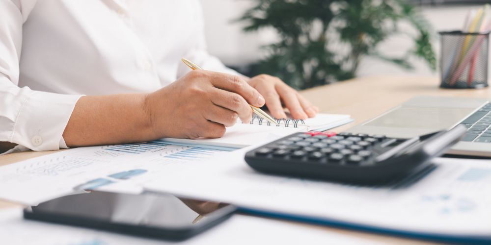 Persona tomando notas junto a una calculadora, documentos y un computador.