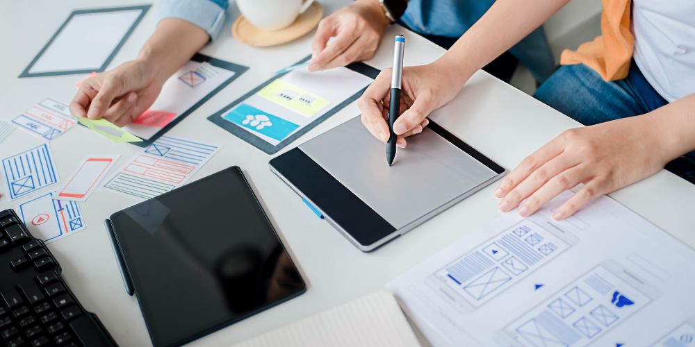 Personas trabajando con una tableta gráfica y bocetos de páginas web sobre una mesa.