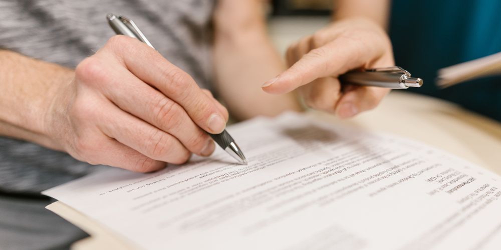 Una persona firma un documento mientras otra señala dónde completar la información.