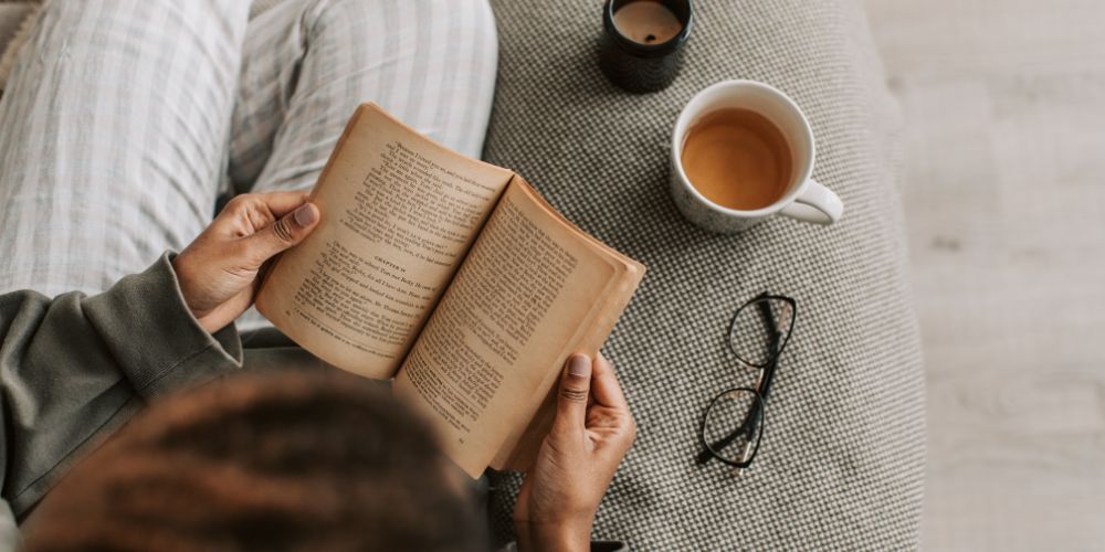 Persona leyendo un libro en un sofá junto a una taza y unos lentes.