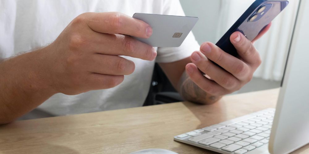 Persona sosteniendo una tarjeta bancaria y un teléfono móvil junto a un computador.