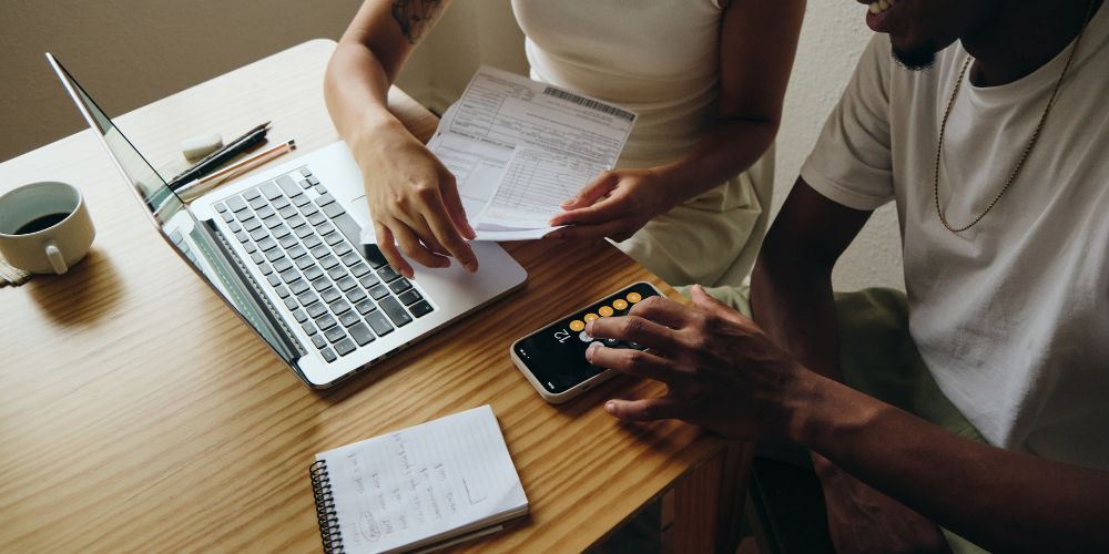 Dos personas revisando documentos y calculando un presupuesto con un teléfono y un computador.