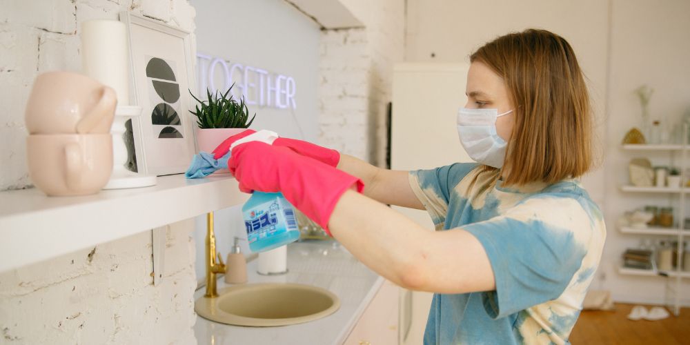 Mujer con guantes y mascarilla limpiando una repisa con un paño y un pulverizador.