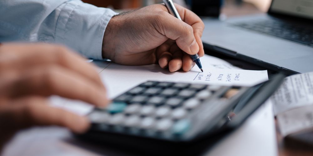 Persona revisando cuentas con calculadora y anotaciones junto a un computador.